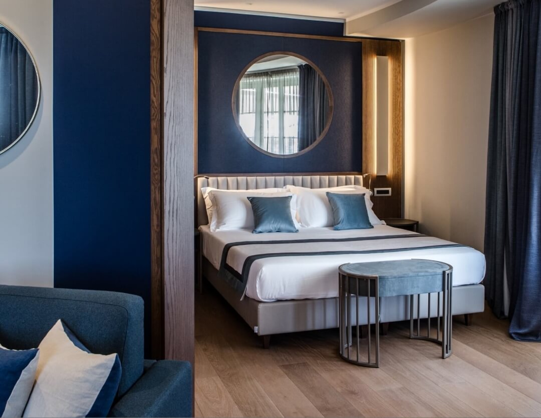 camera da letto alberghiera sulle note del blu notte con lenzuola bianche a contrasto e specchio rotondo sopra la spalliera