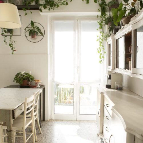 porzione di cucina con tavolo e mobiletto bianchi in legno con piante verdi decorative a contrasto