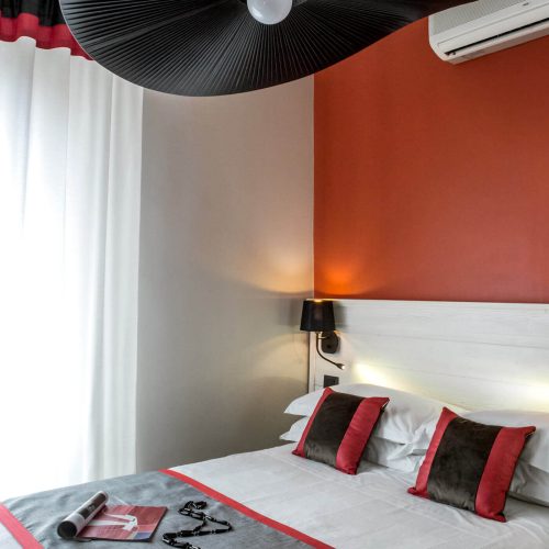 piccola camera d’hotel di design, bianca e rossa con ampio lampadario reazlizzato appositamente evocativo di ampio cappello a falda nero